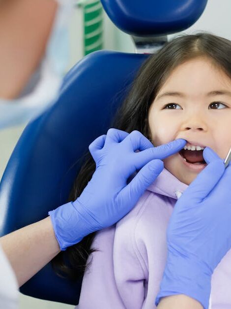 A young girl receives a dental checkup by a dentist wearing gloves in a clinic setting.